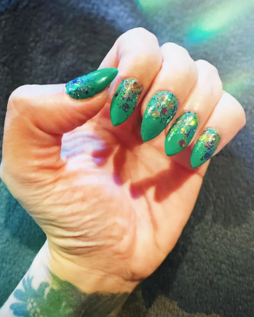 Christmas glitter ombre nails featuring emerald green almond-shaped nails with multicolour gold, blue, and purple glitter concentrated near the cuticles.