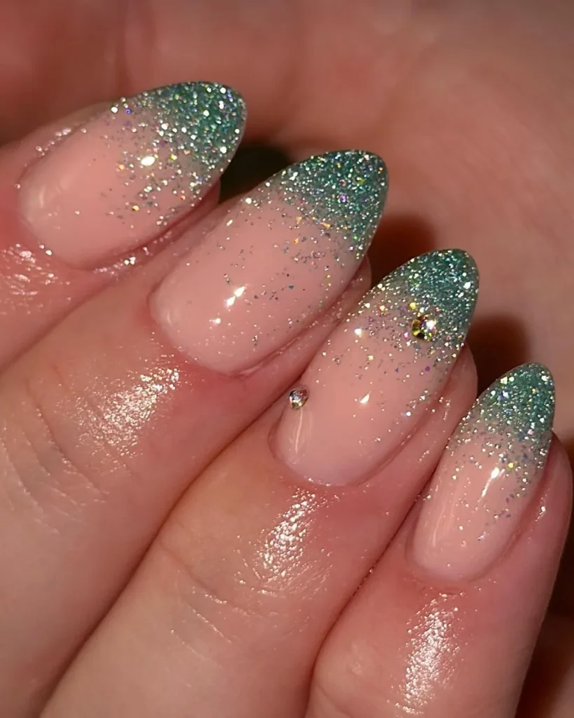 Green Christmas French tip nail designs with emerald green glitter tips, a sheer pink nude base, scattered silver holographic glitter, and a tiny rhinestone accent.