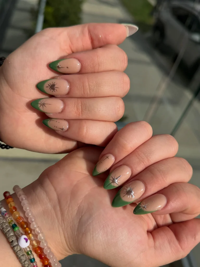 Green Christmas French tip nail designs with deep green almond-shaped tips, a nude beige base, and delicate metallic starburst accents.