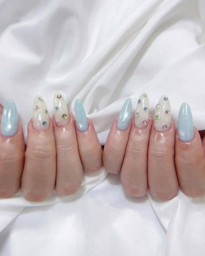 Light blue Christmas nail designs with glossy pastel blue polish, colorful rhinestones, and delicate star accents on long almond-shaped nails.