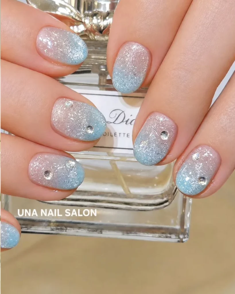 Light blue Christmas nail designs with a pink-to-blue ombre, delicate silver glitter, textured snowflake patterns, and sparkling rhinestone accents on short rounded nails.