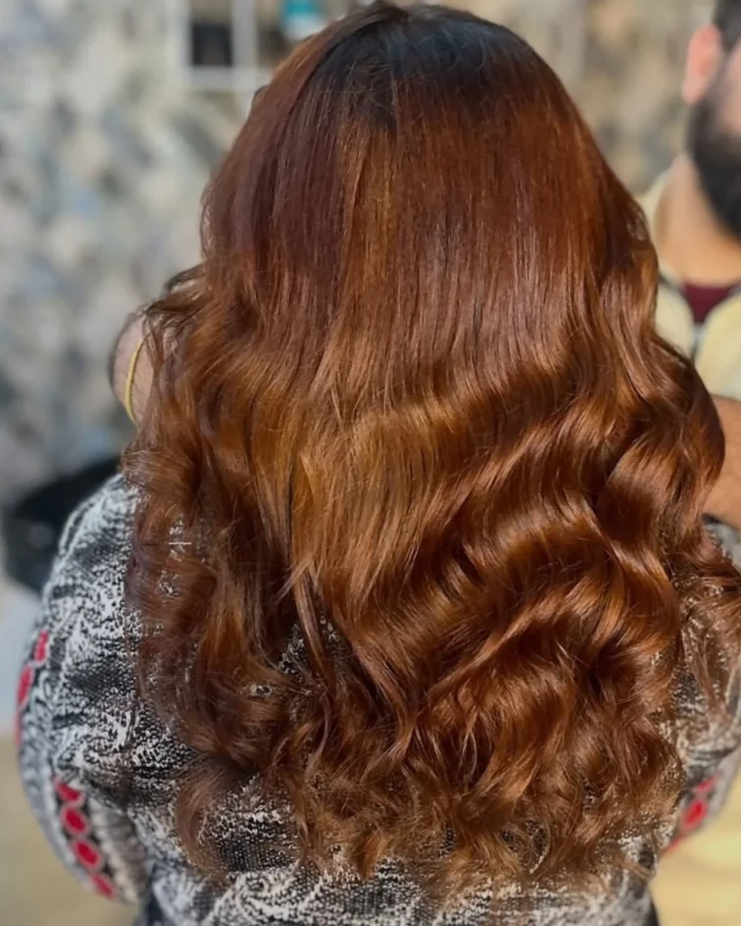 Light golden brown hair color with warm honey-brown and coppery-golden tones styled in long, glossy waves.