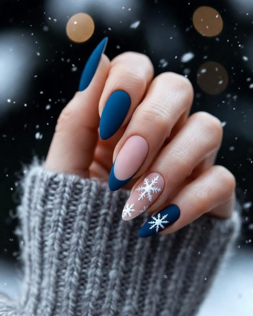 Navy blue Christmas nail designs with white snowflakes, matte polish, nude pink accents, and a French tip on long almond-shaped nails.