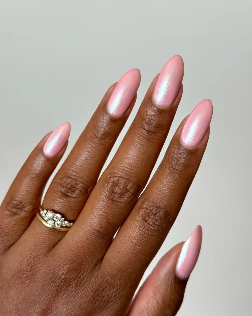 Almond-shaped light pink nails with a pearlescent glazed shimmer and icy reflective highlights on a hand wearing a gold ring.