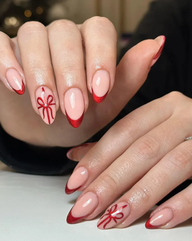 Red French tip Christmas nail designs featuring glittery red tips, glossy nude pink almond-shaped nails, and delicate red gift bow nail art.