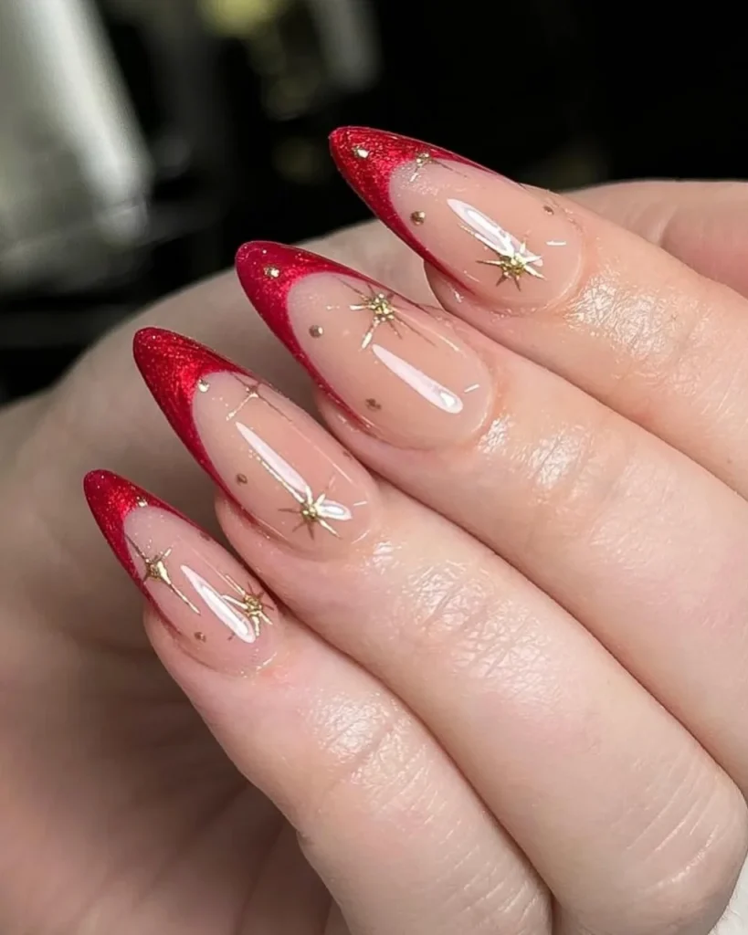 Red French tip Christmas nail designs featuring glittery red tips, glossy nude pink almond-shaped nails, and delicate gold starburst accents for an elegant festive manicure.
