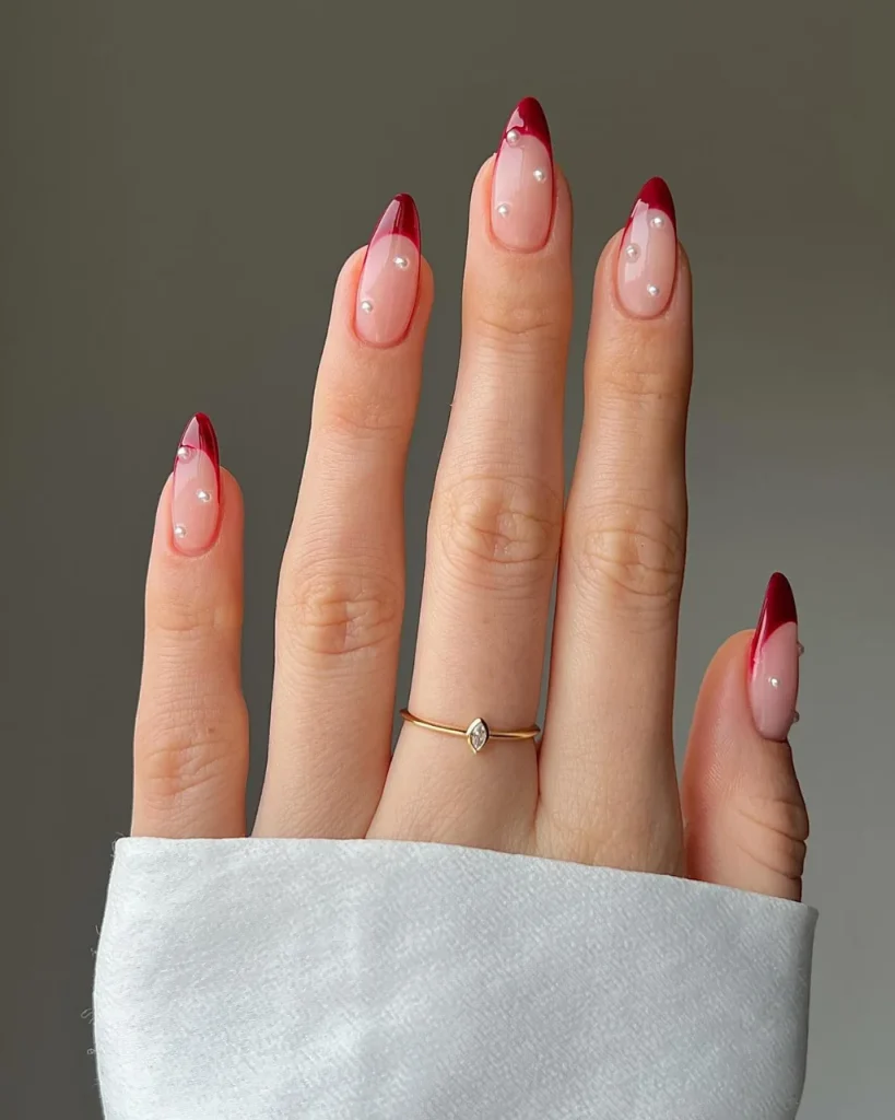 Red French tip Christmas nail designs with glossy burgundy tips, delicate pearl accents, and a sheer pink base on almond-shaped nails.