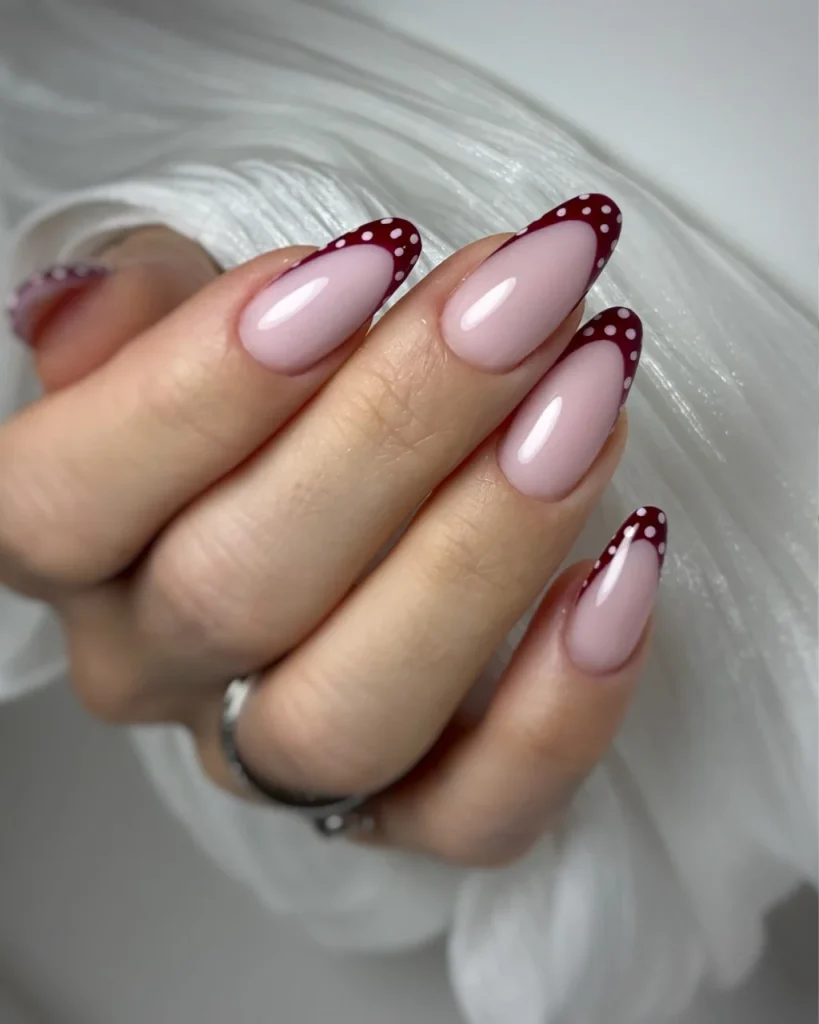 Red French tip Christmas nail designs featuring glossy pink almond-shaped nails with burgundy tips and delicate white polka dots for an elegant festive look.