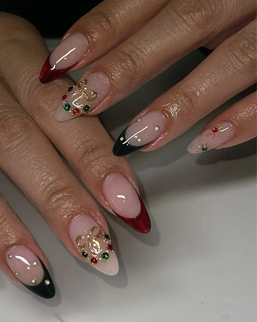 Red French tips with rhinestones, gold bows, and colorful Christmas gems on nude almond-shaped nails.