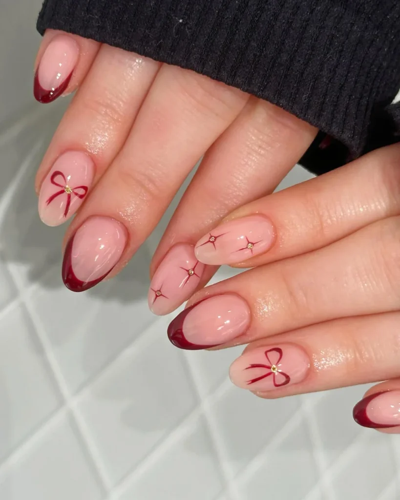 Red French Tips With Tiny Christmas Bows on glossy nude-pink nails with gold accents and delicate starburst details.