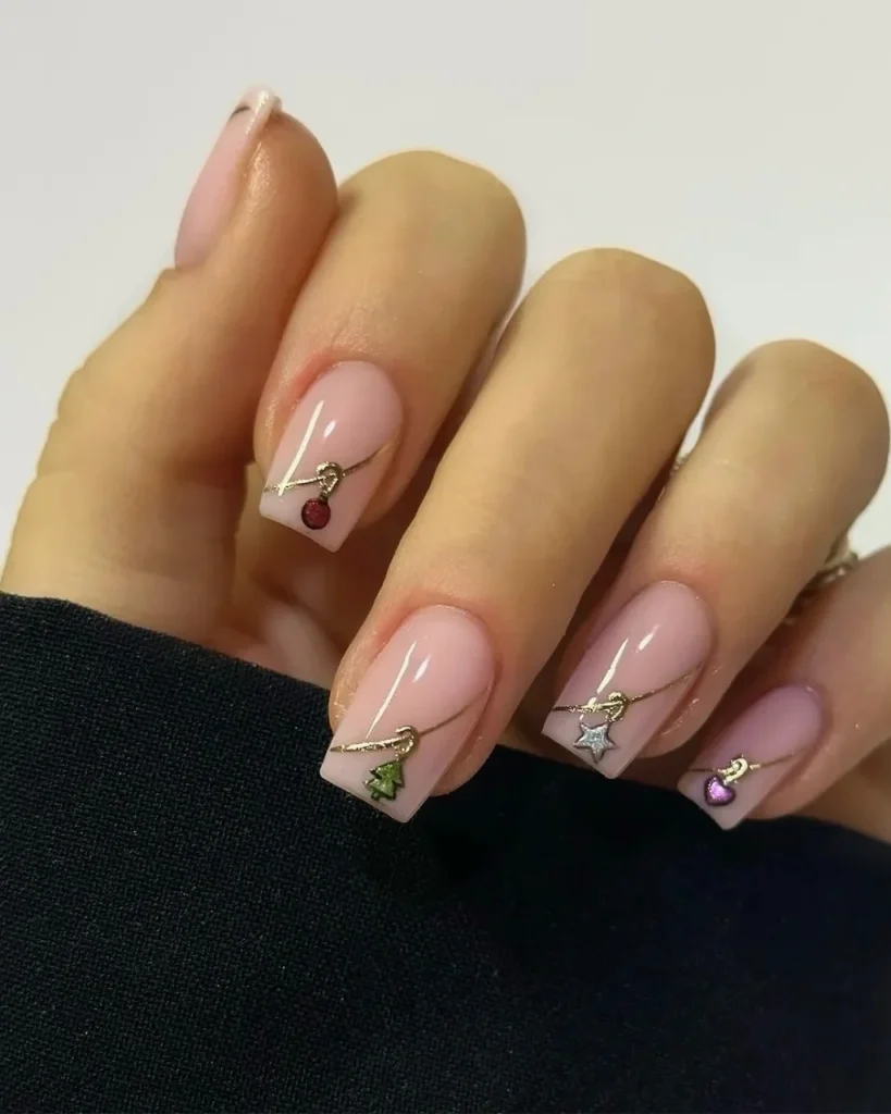 Short Christmas press-on nail designs with a glossy blush-pink base, delicate gold lines, and tiny festive tree, star, cherry, and heart charms.