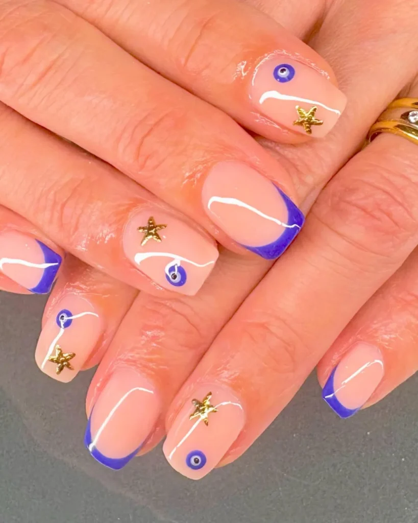 Short french tip christmas nail designs with a glossy peachy nude base, royal blue French tips, white curved lines, and gold star accents.