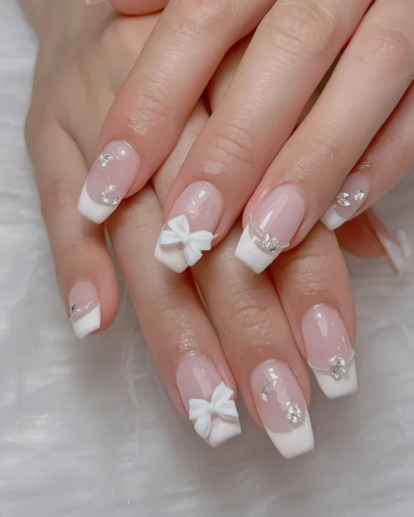 White french tip christmas nail designs with a sheer pink base, 3D white bows, silver rhinestones, and glossy square-shaped nails.