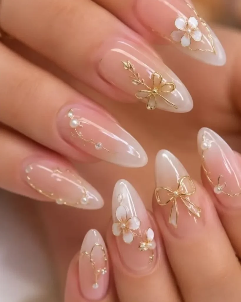 White french tip christmas nail designs with sheer pink almond nails, delicate gold floral art, pearl accents, and elegant bow details.
