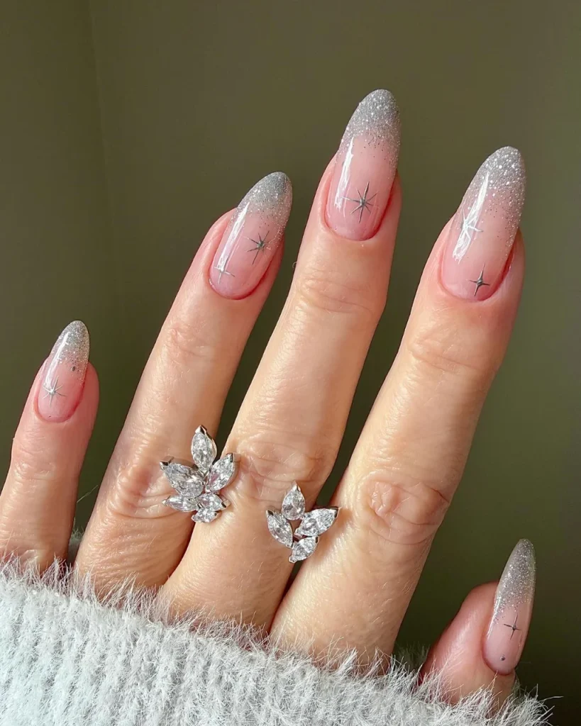 White french tip christmas nail designs featuring silver glitter tips, delicate star accents, and a sheer pink base on almond-shaped nails.